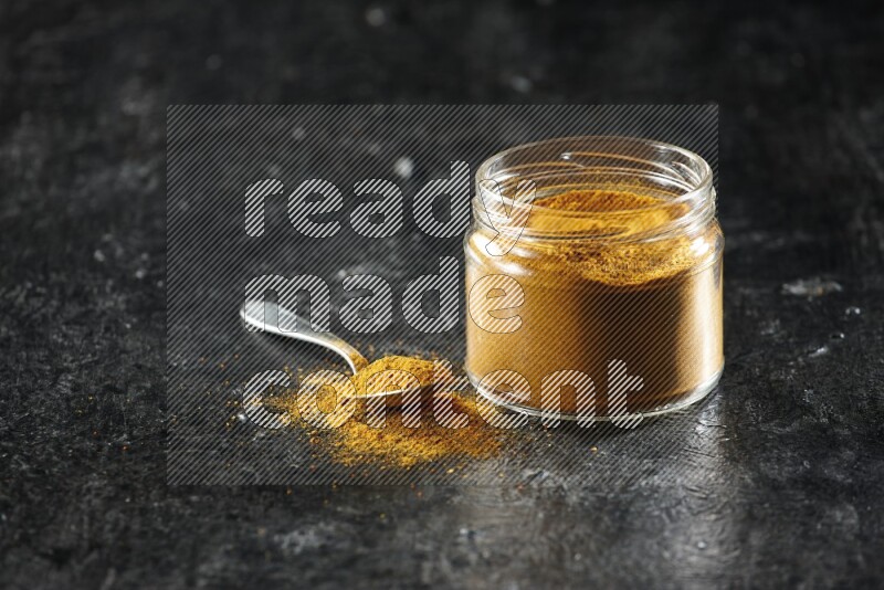 A glass jar and a metal spoon full of turmeric powder on a textured black flooring