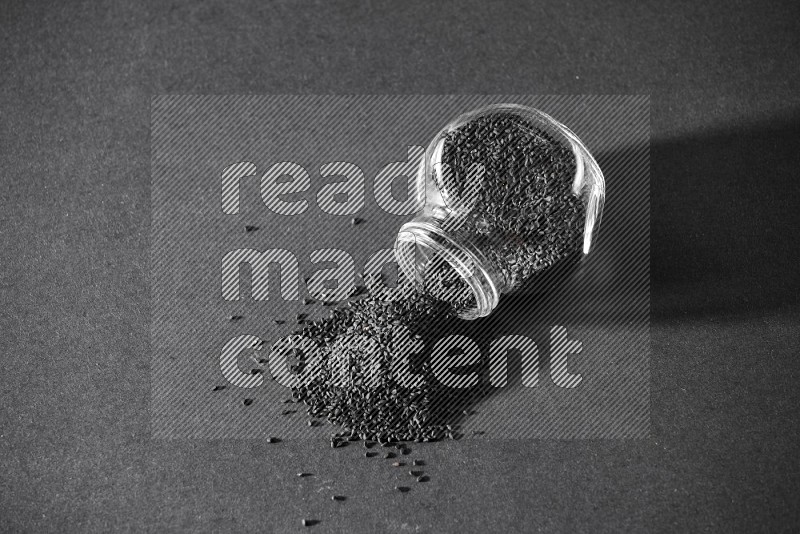 A flipped glass spice jar full of black seeds and the seeds spread out on a black flooring