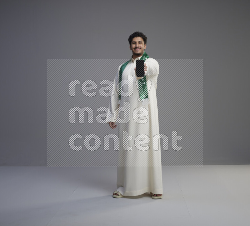 A Saudi man standing wearing thob and Saudi flag scarf showing phone to camera on gray background
