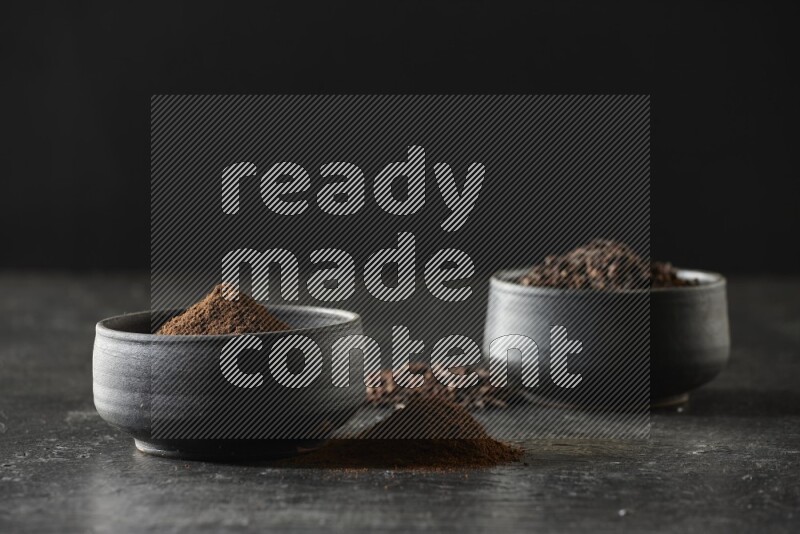 2 Black pottery bowls full of cloves and the other full of cloves powder on textured black flooring