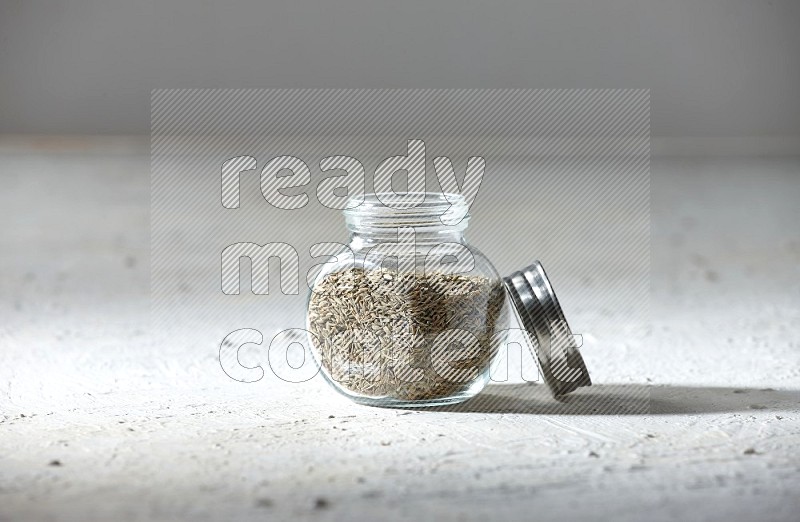 A glass spice jar full of cumin seeds on textured white flooring