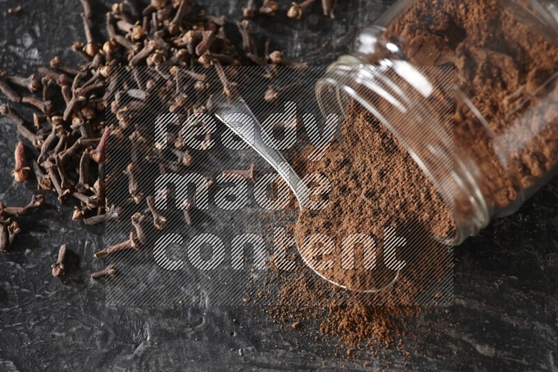 A flipped glass jar and metal spoon full of cloves powder with cloves spread on a textured black flooring