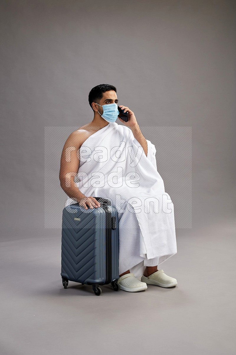 A man wearing Ehram sitting on chair holding traveling bag on gray background