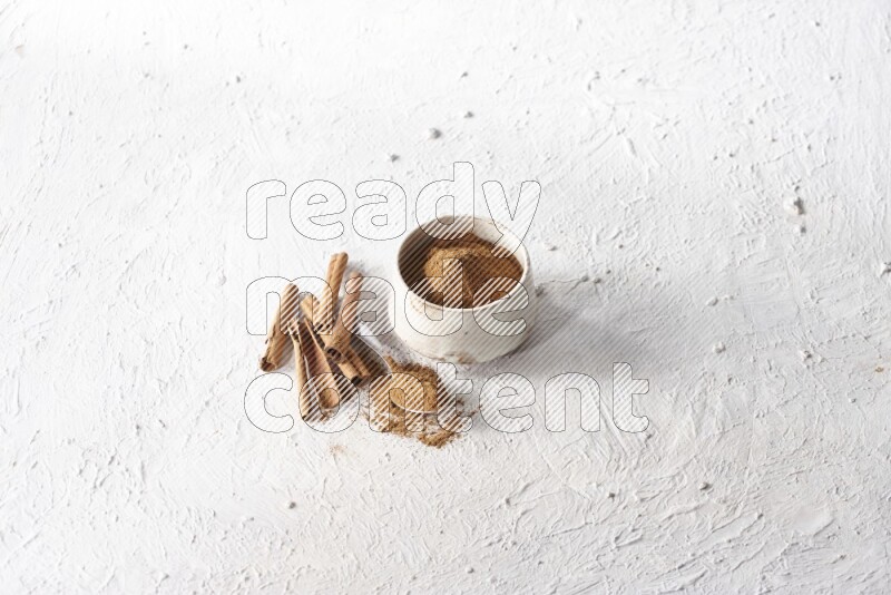 Ceramic beige bowl full of cinnamon powder and a metal spoon with cinnamon sticks next of it on a textured white background