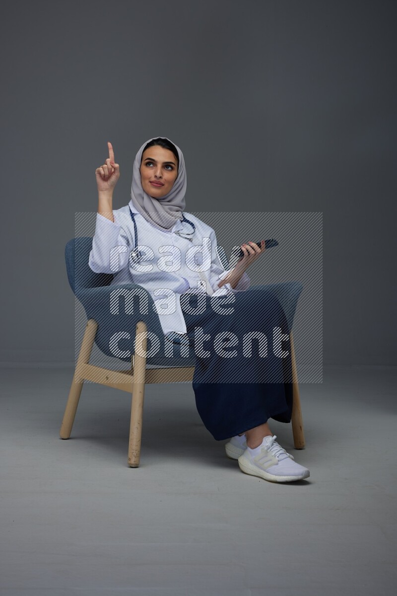 A doctor wearing a light gray head scarf setting on a dark grey chair and using a phone eye level on a grey background