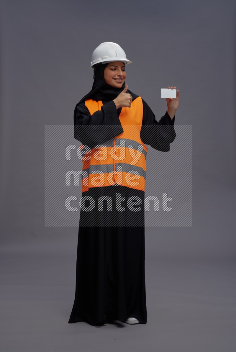 Saudi woman wearing Abaya with engineer vest standing holding ATM card on gray background