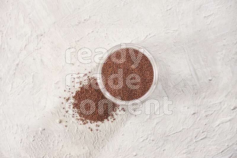 A glass jar full of garden cress seeds with more seeds spread on a textured white flooring