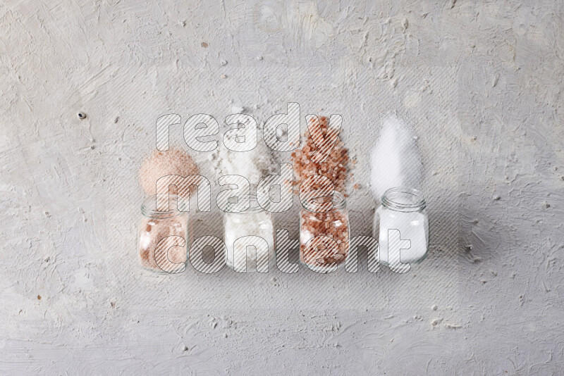 4 glass jars filled with table salt, coarse sea salt, fine himalayan salt and coarse himalayan salt on white background