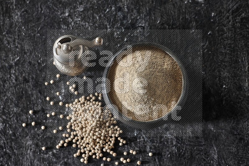 Black pottery bowl full of white pepper powder with pepper beads and metal pepper grinder on textured black flooring