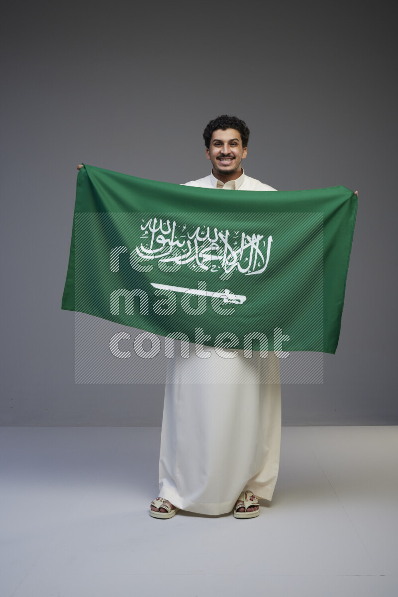 A Saudi man standing wearing thob holding big Saudi flag on gray background