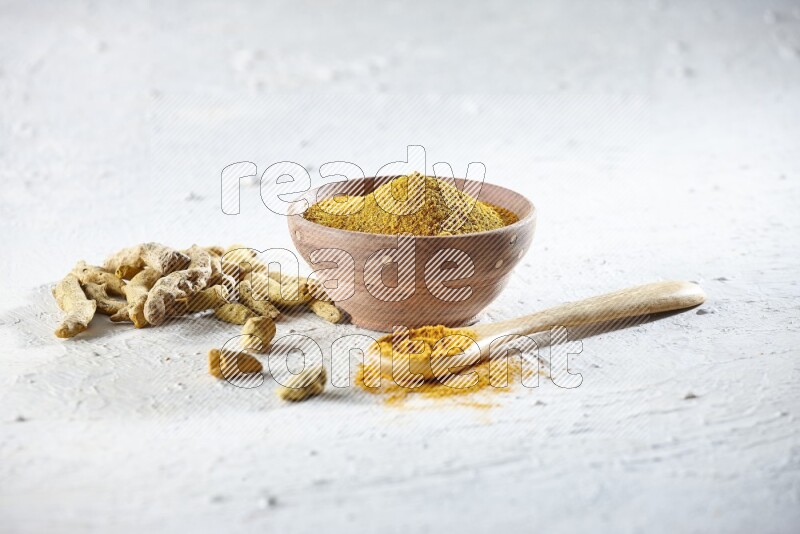 A wooden bowl and wooden spoon full of turmeric powder with dried turmeric fingers beside it on textured white flooring