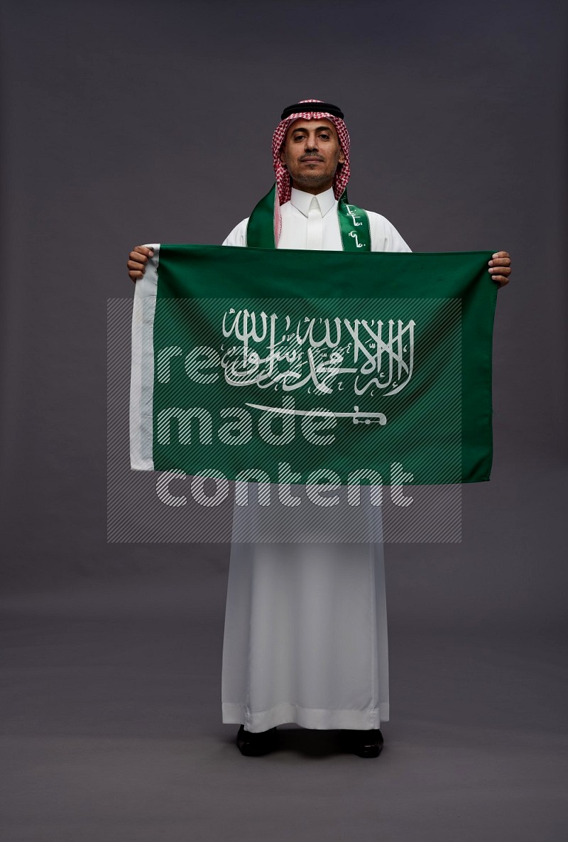 Saudi man wearing thob and shomag standing holding Saudi flag on gray background