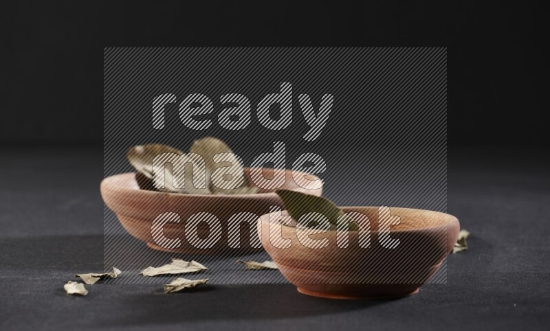 2 wooden bowls full of dried bay leaves with more leaves spread on black flooring