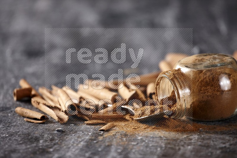 Herbal glass jar full cinnamon powder flipped and a metal spoon full of powder surrounded by cinnamon sticks on textured black background in different angles