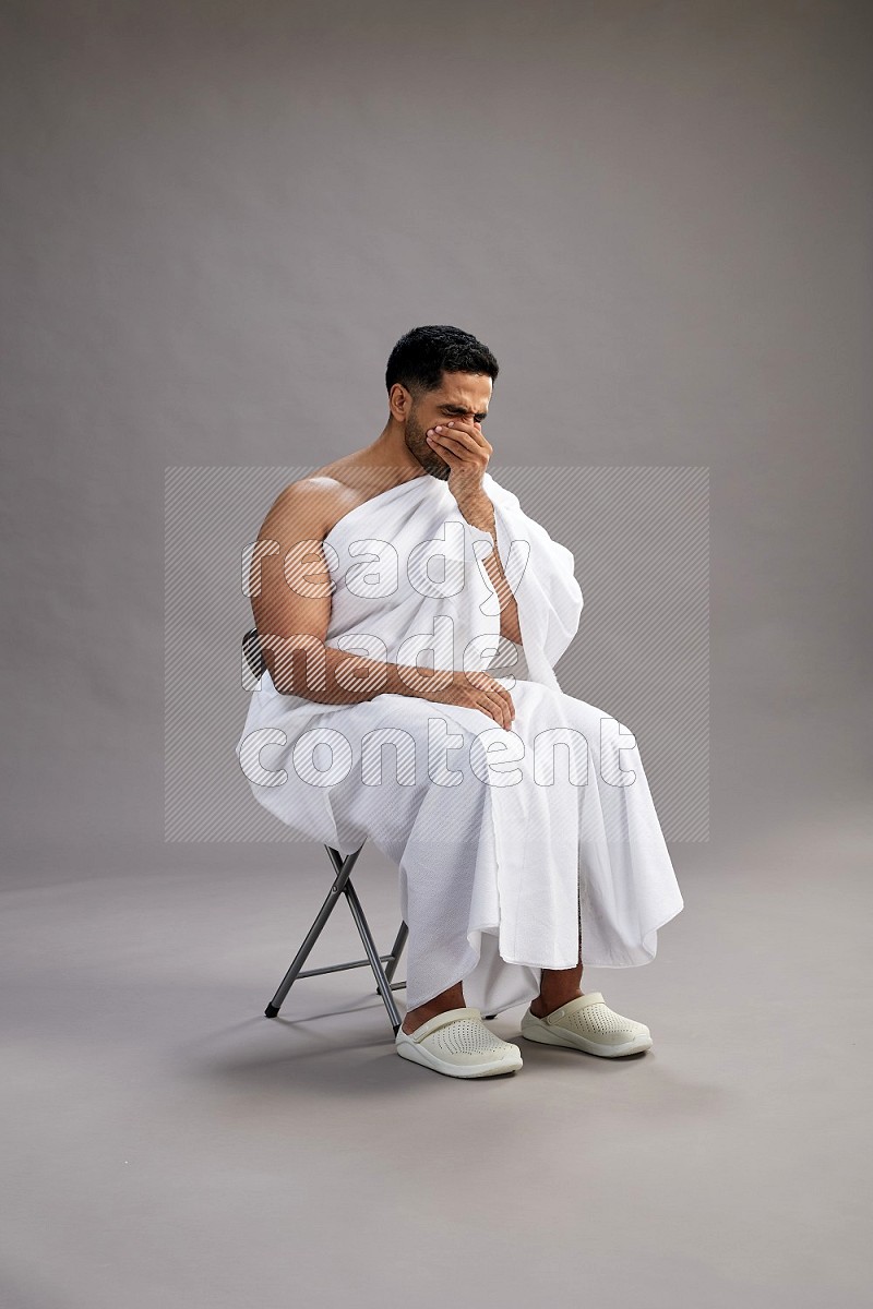 A man wearing Ehram sitting on chair showing how to sneeze on gray background