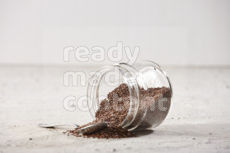 A glass spice jar full of flax seeds flipped with a metal spoon full of the seeds on a textured white flooring