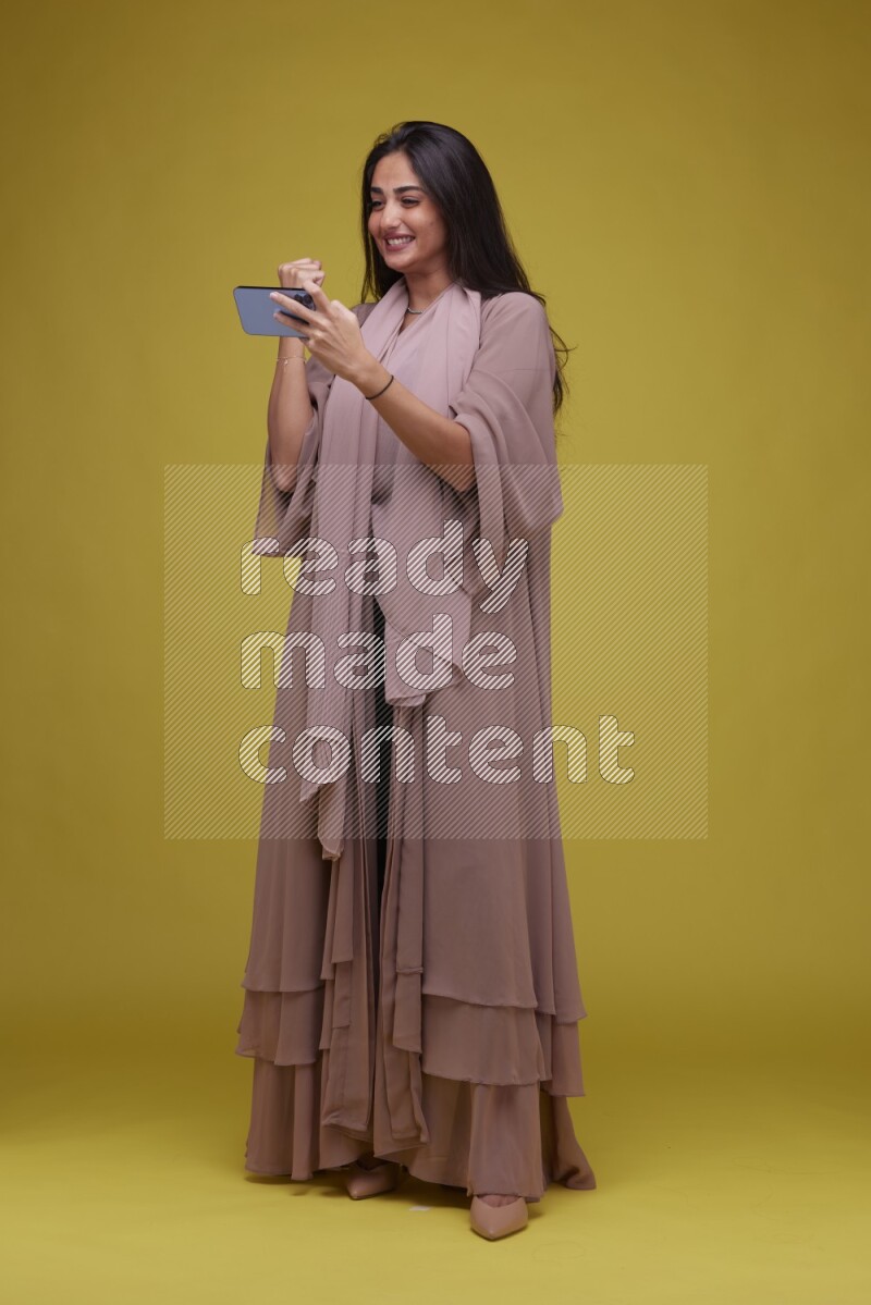 A woman Playing a Game on her smartphone on a Yellow Background wearing Brown Abaya