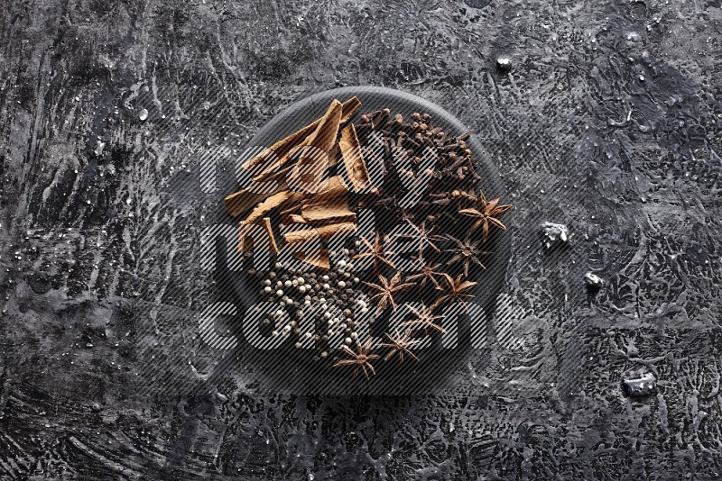 Cinnamon sticks, cloves, star anise and black and white peppers on a black plate on textured black background