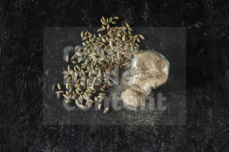 A flipped glass spice jar and a metal spoon full of cardamom powder and cardamom seeds spreaded on textured black flooring