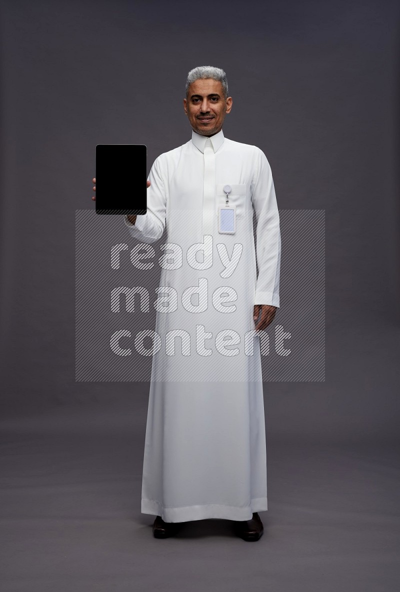 Saudi man wearing thob with pocket employee badge standing showing tablet to camera on gray background