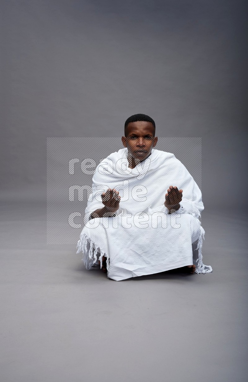 A man wearing Ehram sitting on floor dua'a on gray background