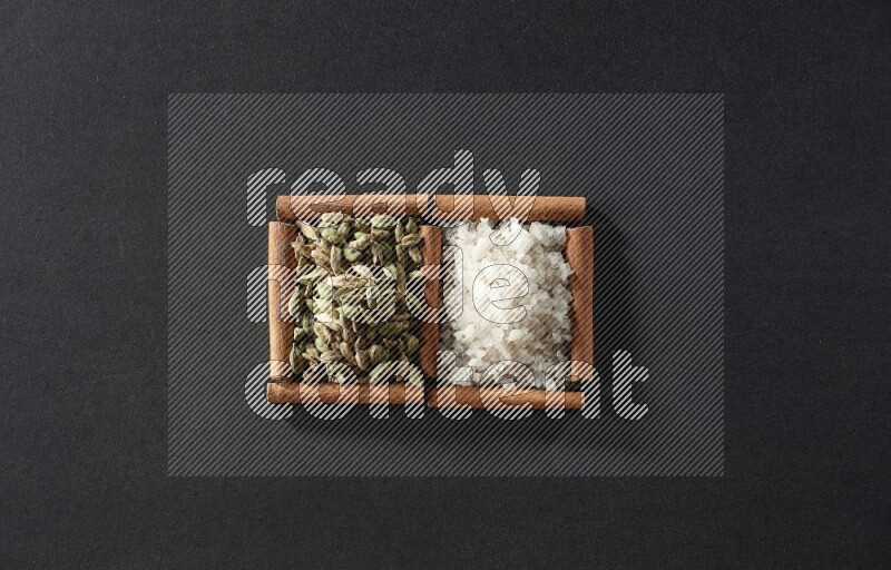 مربعان من أعواد القرفة ممتلئان بالملح الأبيض وحبوب الهيل علي خلفية سوداء