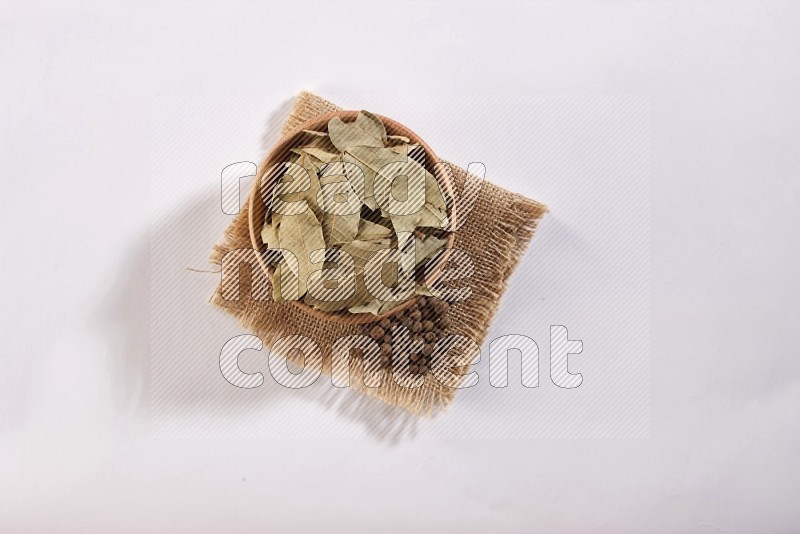A wooden bowl filled with dried bay leaves on a piece of burlap with some of allspice berries on white flooring in different angles