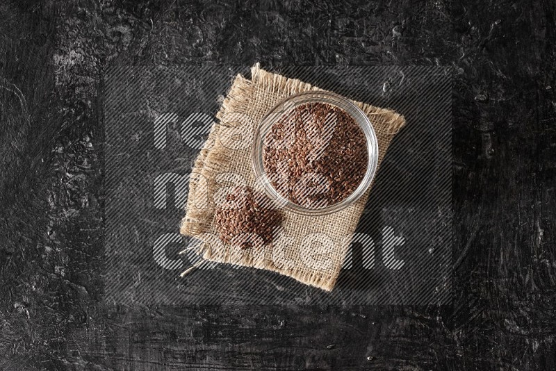 A glass bowl full of flaxseeds with bunch of the seeds on burlap fabric on a textured black flooring