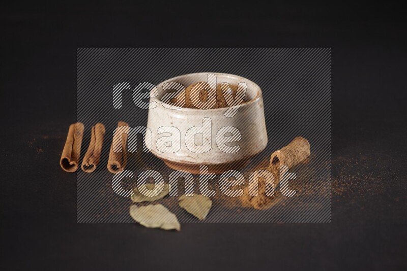 Cinnamon powder in a white pottery bowl and cinnamon sticks and laurel leaves on black background