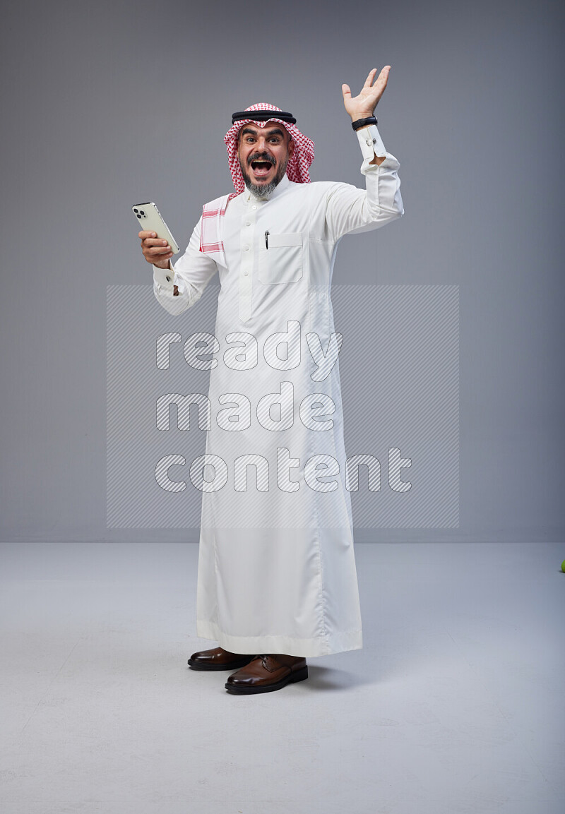 Saudi man Wearing Thob and red Shomag standing texting on phone on Gray background