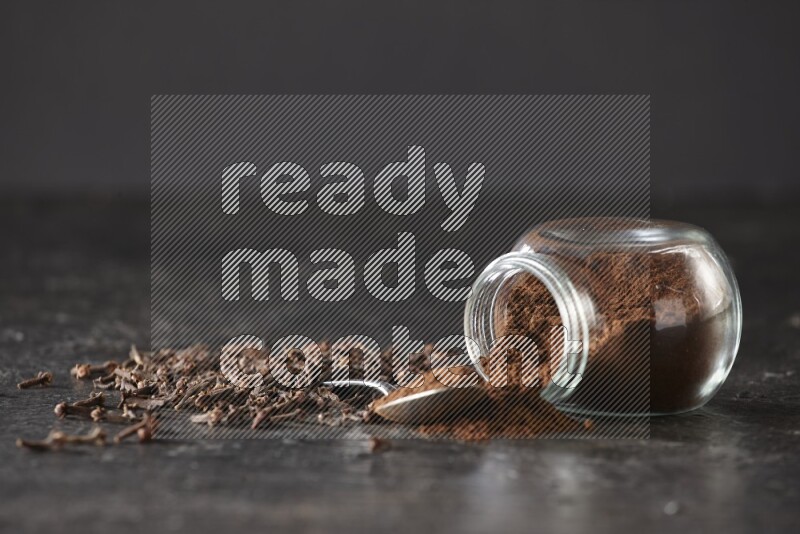 A flipped glass spice jar and a metal spoon full of cloves powder with cloves spread on textured black flooring