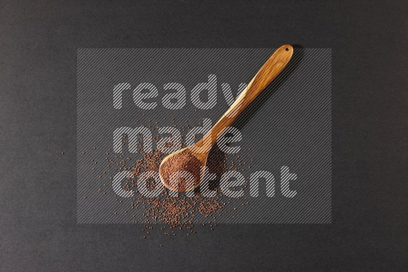 A wooden ladle full of garden cress seeds and seeds spread beside it on a black flooring