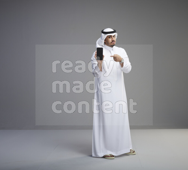 A Saudi man standing wearing thob and white shomag showing phone to camera on gray background