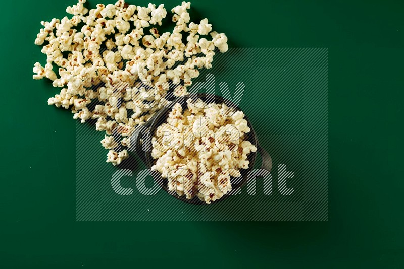 A black ceramic bowl full of popcorn with popcorn beside it on a green background in a top view shot