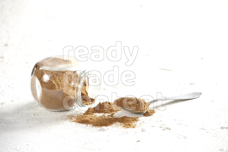 Flipped herbs glass jar full of cinnamon powder with a metal spoon full of powder on a textured white background