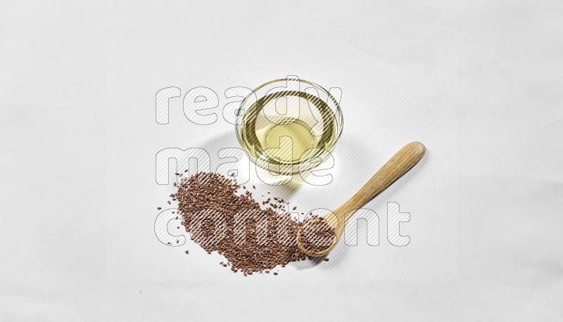A glass bowl full of flaxseeds oil and wooden spoon full of flaxseeds with spread seeds on a white flooring