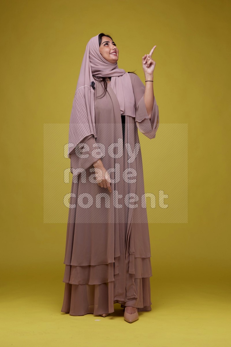 A woman Pointing on a Yellow Background wearing Brown Abaya with Hijab