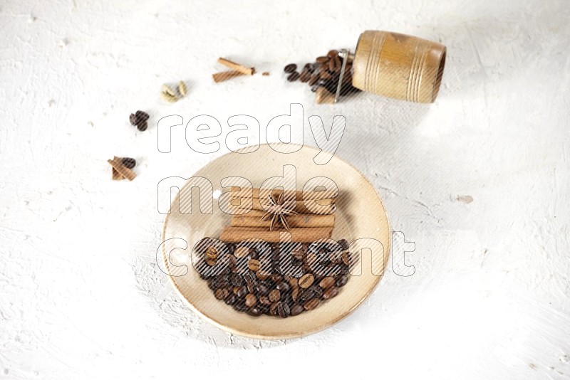 Beige plate full of coffee beans, cinnamon sticks and star anise with a coffee grinder, coffee beans, cinnamon pieces and cardamom next of it on white background