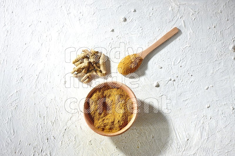 A wooden bowl and wooden spoon full of turmeric powder with dried turmeric fingers on textured white flooring