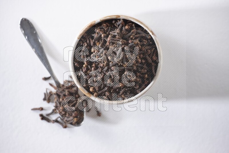 وعاء بيج سيراميك مملوء بالقرنفل وبجانبه ملعقة معدنية على أرضية بيضاء