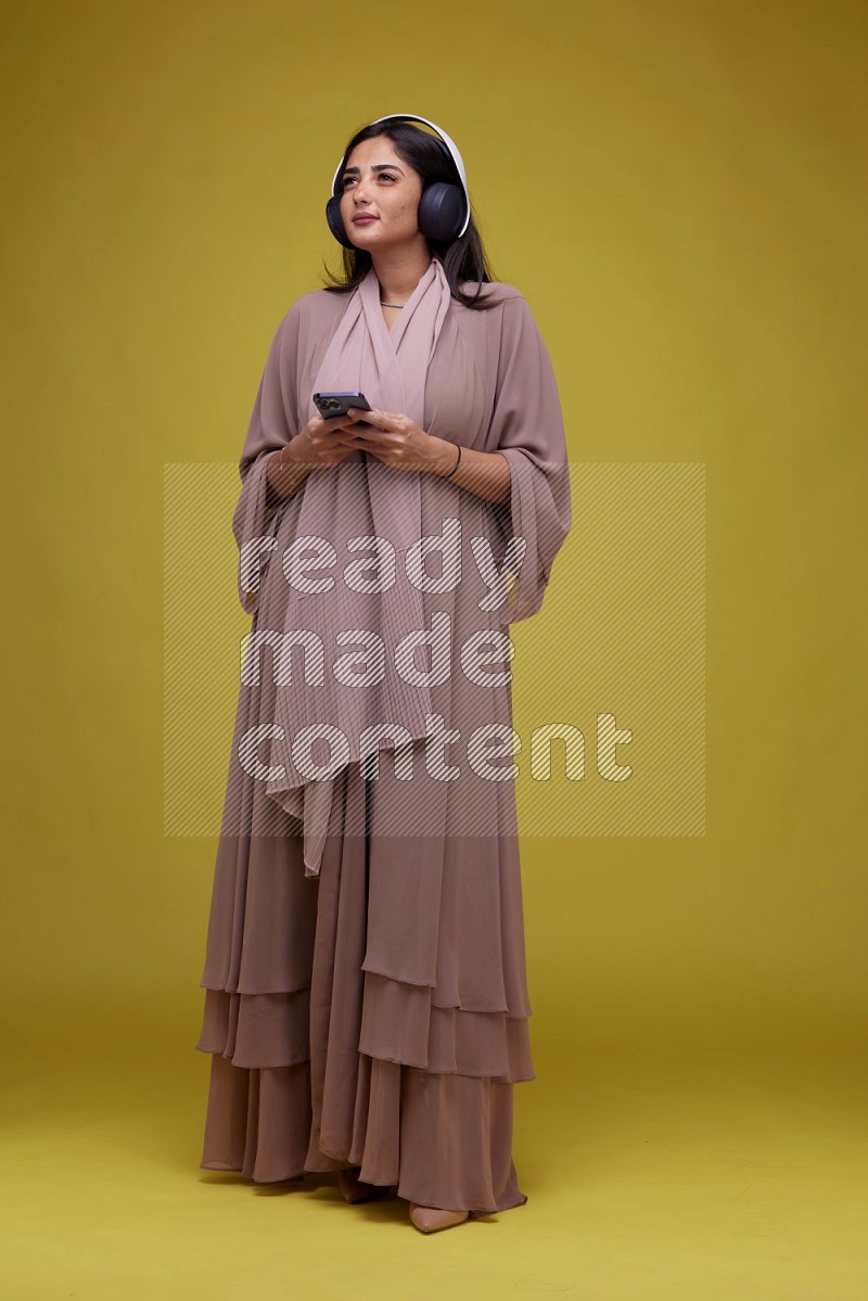 A Saudi woman Listing to Music on a Yellow Background wearing Brown Abaya