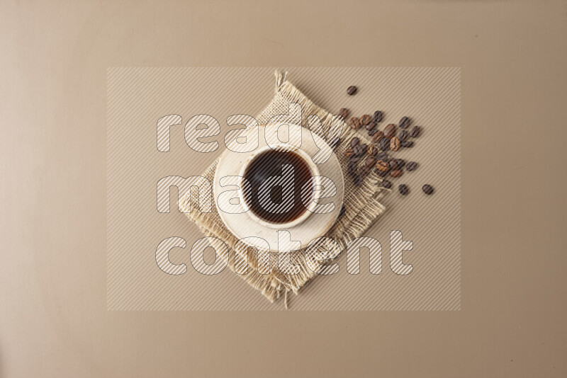 A beige pottery cup of coffee surrounded by roasted coffee beans on beige background