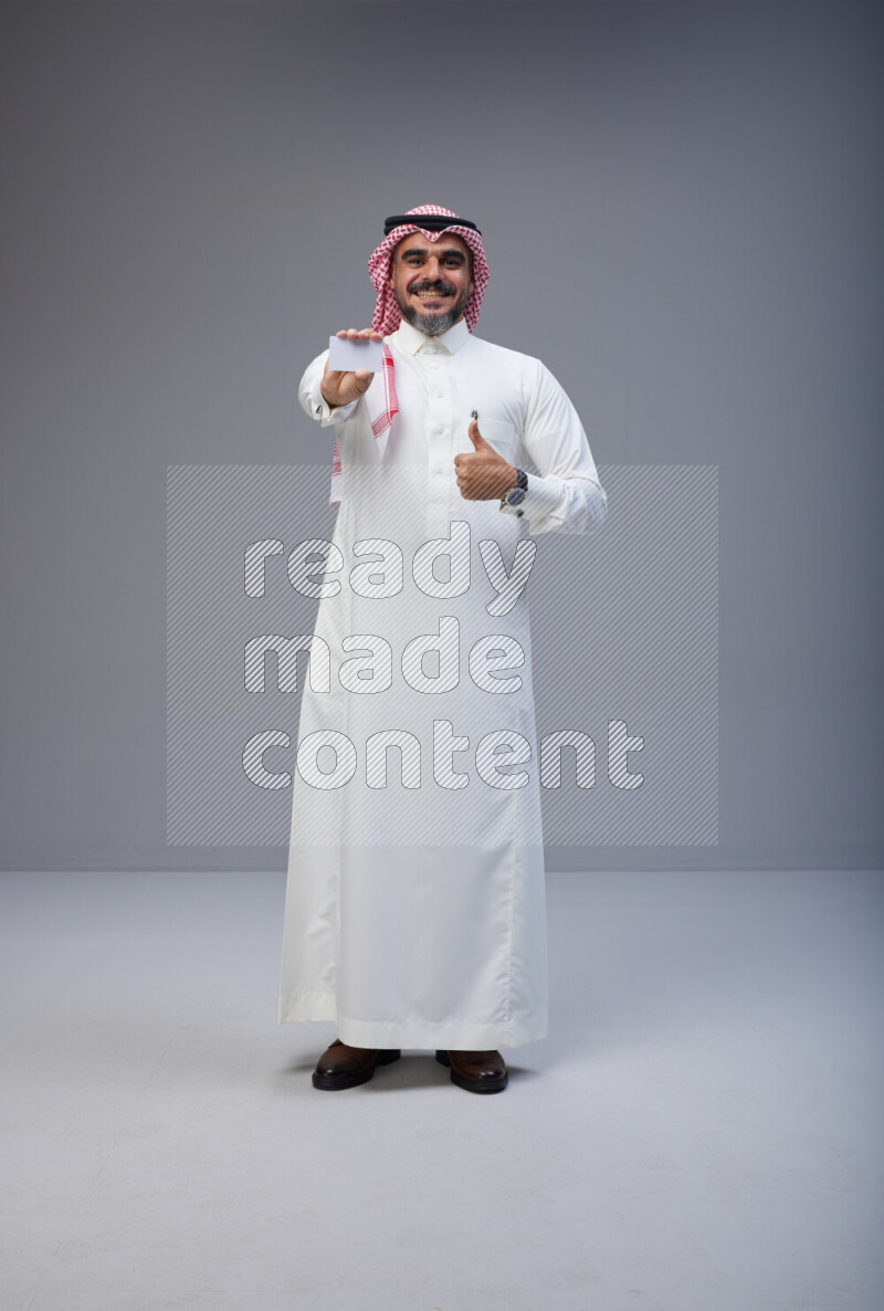 Saudi man Wearing Thob and red Shomag standing holding ATM card on Gray background