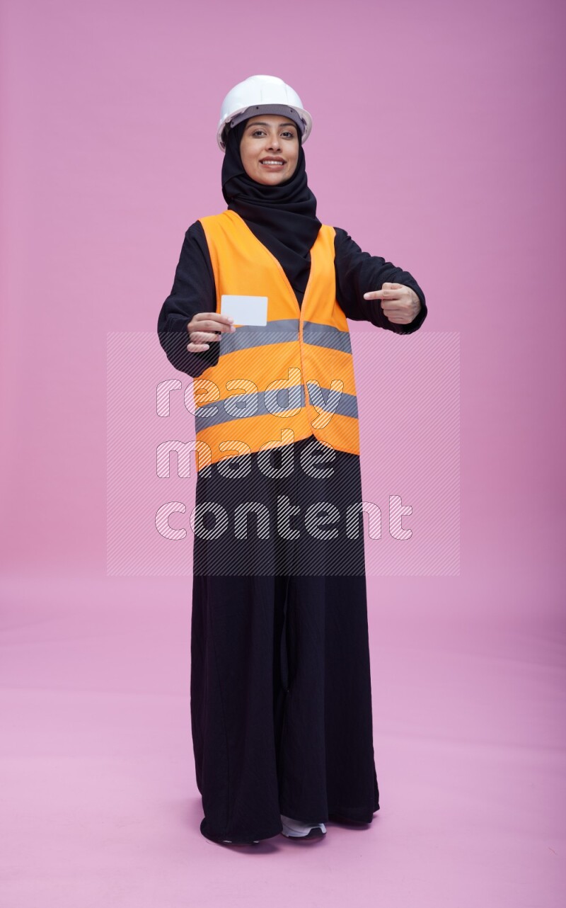 Saudi woman wearing Abaya with engineer vest and helmet standing holding ATM card on pink background