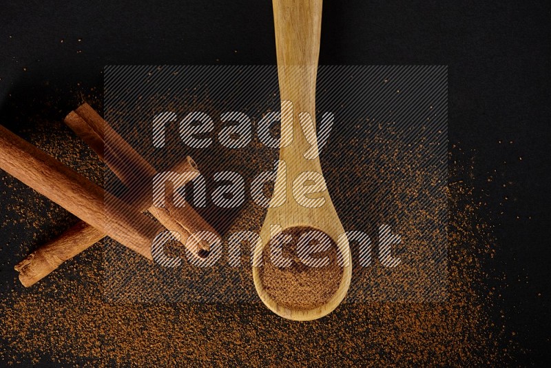 Cinnamon powder in a wooden spoon and cinnamon sticks beside it on black background