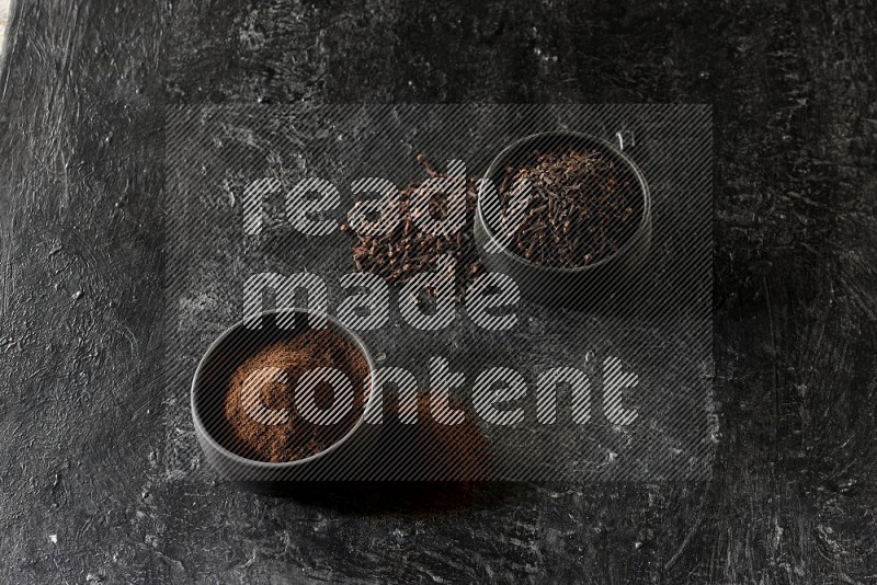 2 Black pottery bowls full of cloves and the other full of cloves powder on textured black flooring