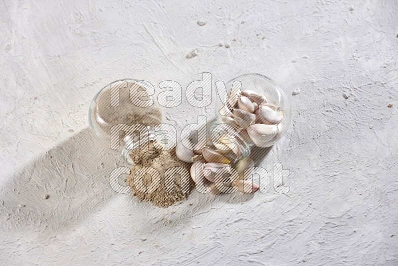2 glass spice jars full of garlic cloves and powder flipped and their garlic came out on a textured white flooring in different angles