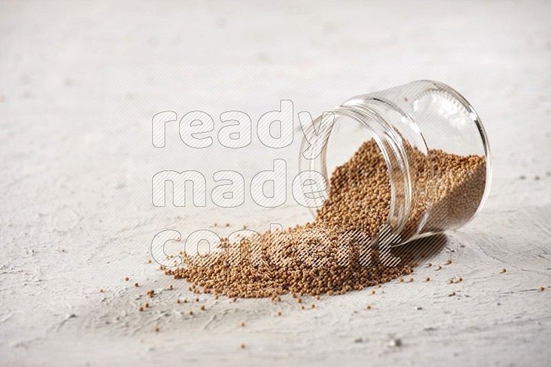A glass jar full of mustard seeds and jar is flipped with seeds spread out on a textured white flooring
