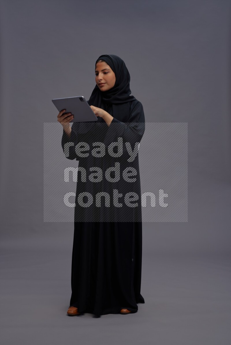 Saudi woman wearing Abaya standing working on tablet on gray background