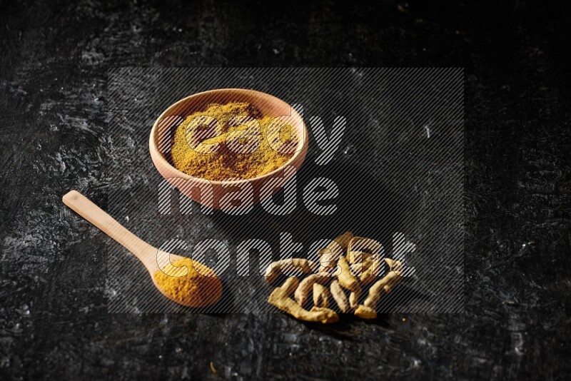 A wooden bowl and a wooden spoon full of turmeric powder with dried turmeric fingers on textured black flooring